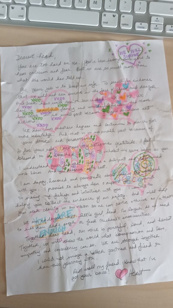 A handwritten letter sits on a desk below a computer keyboard. The paper contains rips and folds and, around the text, there are doodles of hearts made with fluorescent markers.