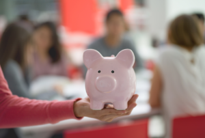 woman's arm, in foreground, holding a pink piggy bank with group of adults sitting, out of focus, in the background.