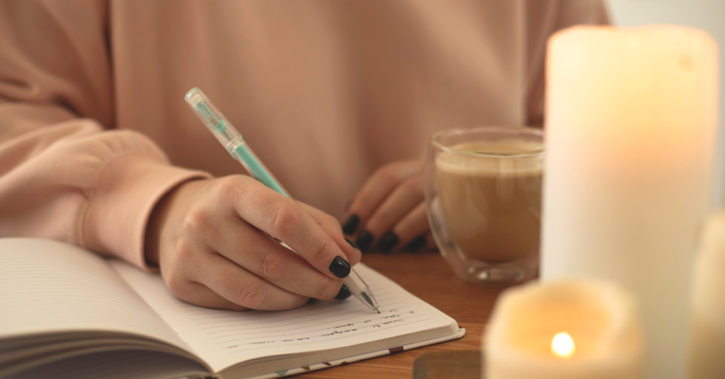 A woman's hand, holding a blue pen, writing in a journal by candle light.