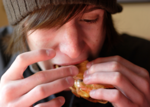 close-up on the face of a teenage boy wearing a gray wool cap and eating a greasy hamburger