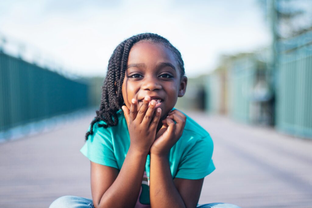 young black girl with braided hair, wearing a teal green shirt, sits on a boardwalk looking at the camera. Photo by Kindred Hues on Unsplash. https://unsplash.com/@kindredhues