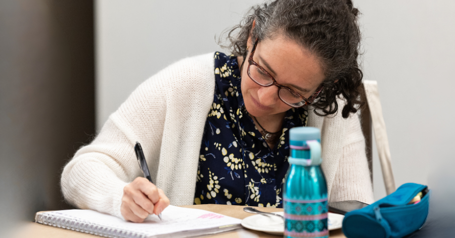 woman with curly hair and glasses wearing a blue dress with yellow flowers and a white sweater is seated at a table writing in a notebook.