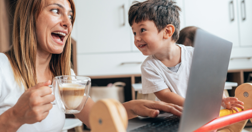 A mom with long blond hair and a cup of coffee sits at the table with her computer and makes a funny face with her young son who is laughing with her.