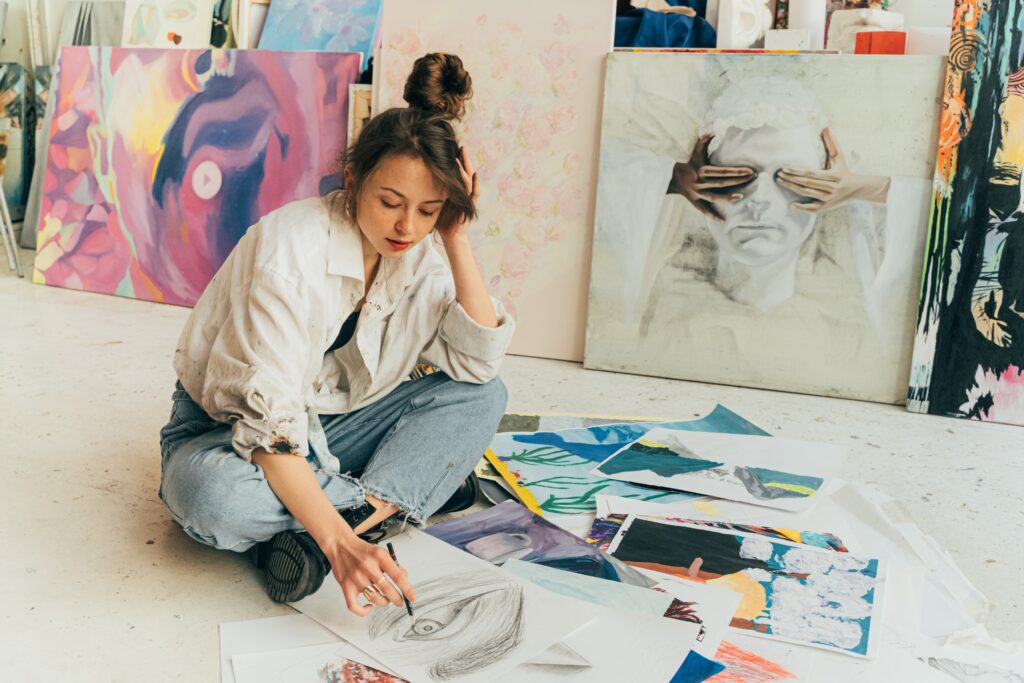 woman with brown hair, wearing a white button-down shirt and jeans, sits on the floor sketching and image of an eye whlle other works of art lay in a pile. Behind her, there are paintings leaning against a wall. Photo by Antoni Shkraba. Downloaded from Pexels.
