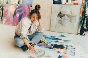 woman with brown hair, wearing a white button-down shirt and jeans, sits on the floor sketching and image of an eye whlle other works of art lay in a pile. Behind her, there are paintings leaning against a wall. Photo by Antoni Shkraba. Downloaded from Pexels.