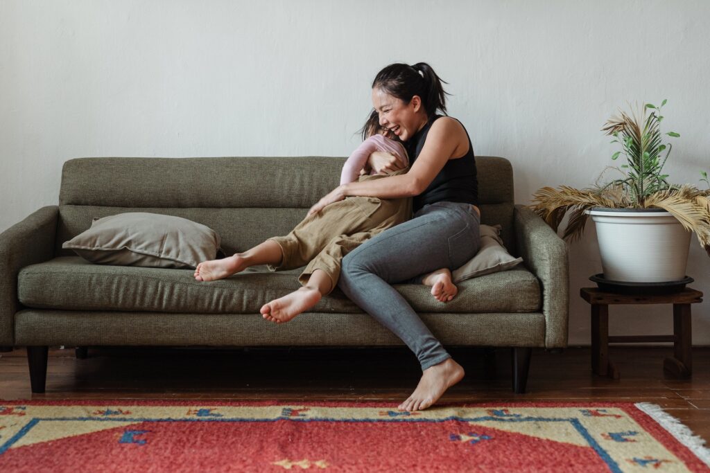 Mother and son sitting on a gray couch in a playful hug. Both are barefoot and laughing.