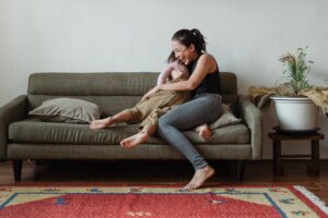 Mother and son sitting on a gray couch in a playful hug. Both are barefoot and laughing.